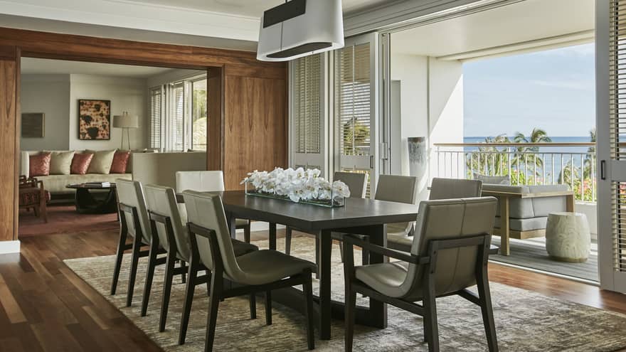 A modern dining room with a large table and elegant chairs, opening to a balcony with ocean views. The space features wood accents, soft lighting and floral decor.