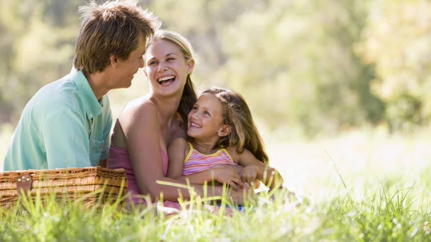 Smiling man, woman and young girl sit in tall grass by picnic basket on sunny day