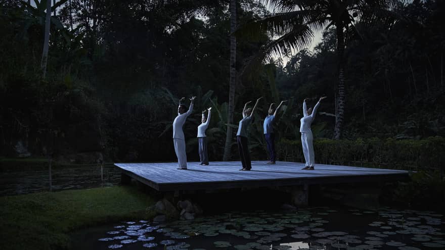 Group of five people participate in full moon Purnama yoga on a floating wooden deck above a pond of lily pads
