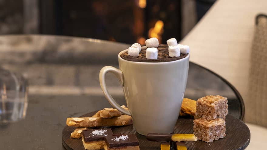 White mug of hot chocolate with mashmallows on a dark, round platter filled with small desserts