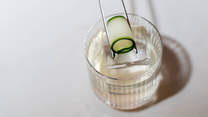 A thin, rolled up slice of cucumber is being placed atop a cocktail by a set of slim silver tongs
