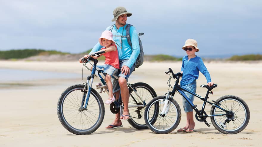 Family on beach, man wearing hat sits on bicycle, young girl in front, boy stands with children's bike