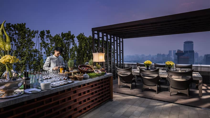 Bartender prepares drinks, cheeses, salads at long Imperial Suite rooftop bar by private dining table at night