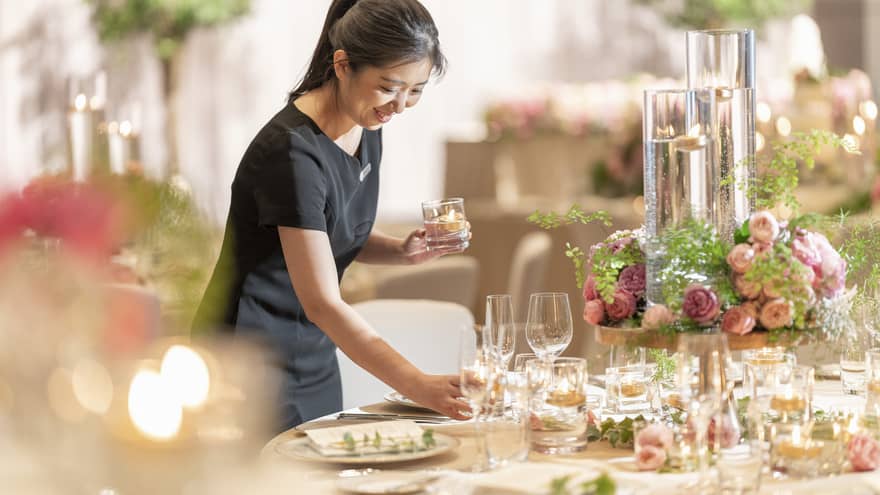 A smiling staff member arranging a table with a floral centerpiece, long glass jars with floating tea lights and champagne glasses