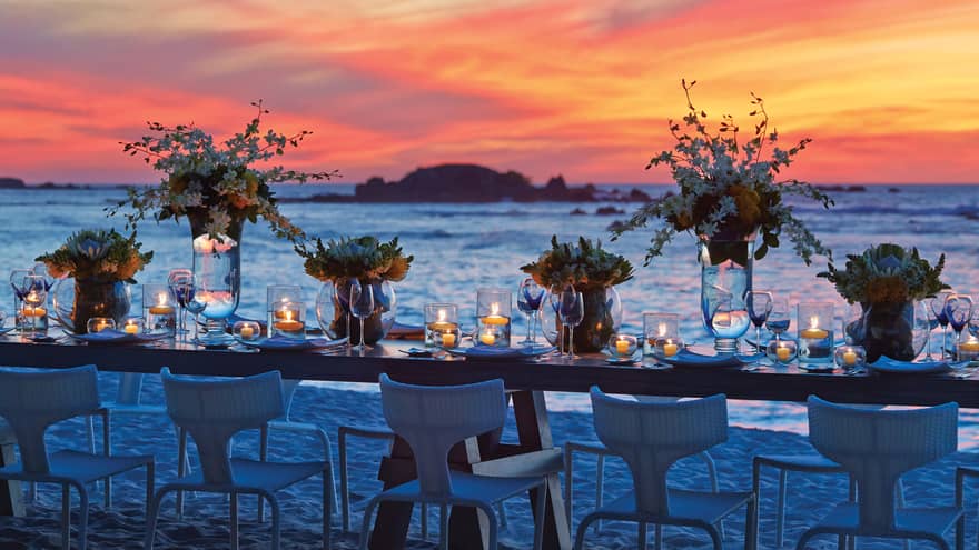 A long candle-lit wedding table with floral decorations on the white sand beach at Punta Mita, set against a vibrant sunset backdrop
