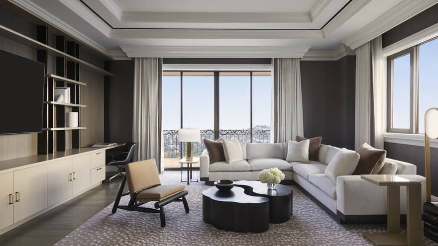 Living room with l-shaped white sofa, two-piece abstract coffee table, chair, wall-mounted TV and wall of windows