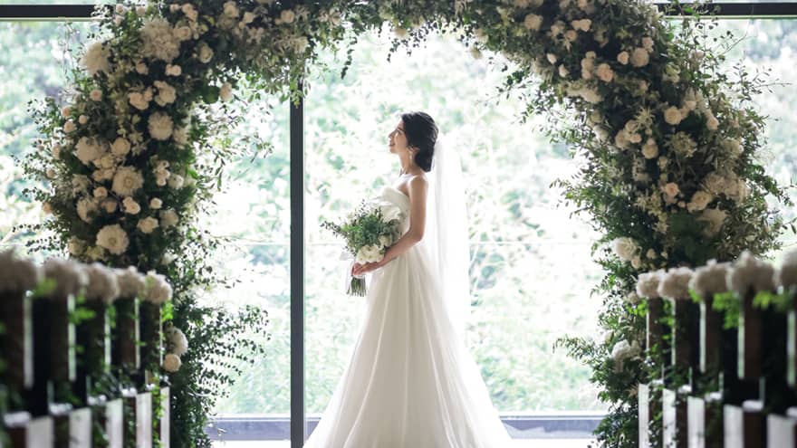 Bride stands under round wedding altar covered in white flowers by sunny window