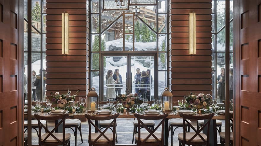 Elegant winter wedding setup inside Four Seasons Whistler, viewed through glass doors leading to a cozy, beautifully decorated dining area. The room is filled with wooden tables set with fine dinnerware, crystal glassware, and lush greenery centerpieces, all illuminated by warm overhead lights. Outside the glass doors, guests in formal attire chat in a snowy courtyard, adding to the festive and romantic atmosphere.,Elegant winter wedding setup inside Four Seasons Whistler, viewed through glass doors leading to a cozy, beautifully decorated dining area. The room is filled with wooden tables set with fine dinnerware, crystal glassware, and lush greenery centerpieces, all illuminated by warm overhead lights. Outside the glass doors, guests in formal attire chat in a snowy courtyard, adding to the festive and romantic atmosphere.
