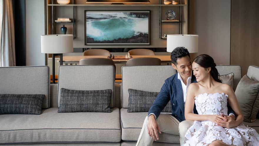 Smiling couple seated on a light gray sofa in a modern living room with decorative shelves and a large ocean artwork in the background.