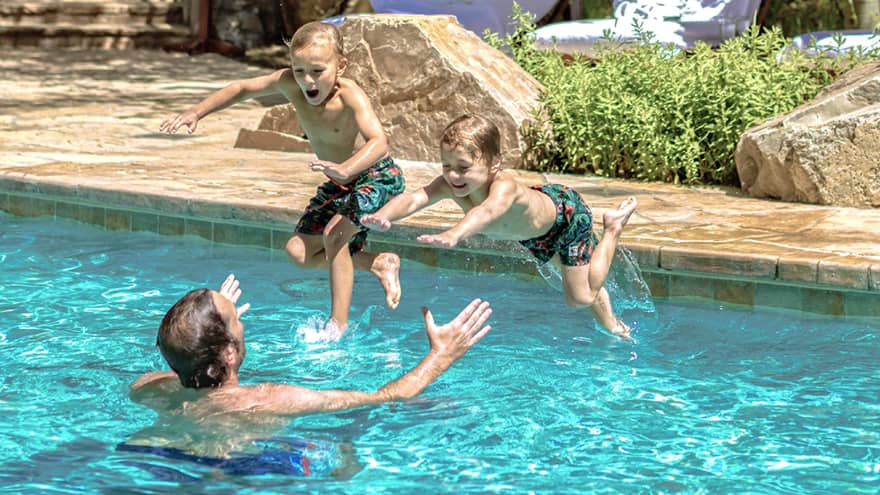 Two young boys jump into their father's outstretched arms in an outdoor pool 