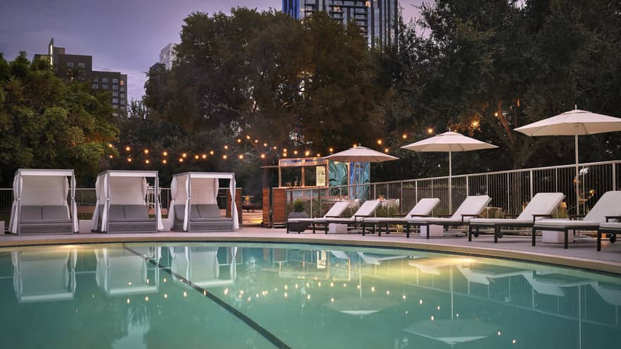 An outdoor pool at evening surrounded by lounge chairs, umbrellas and large trees.