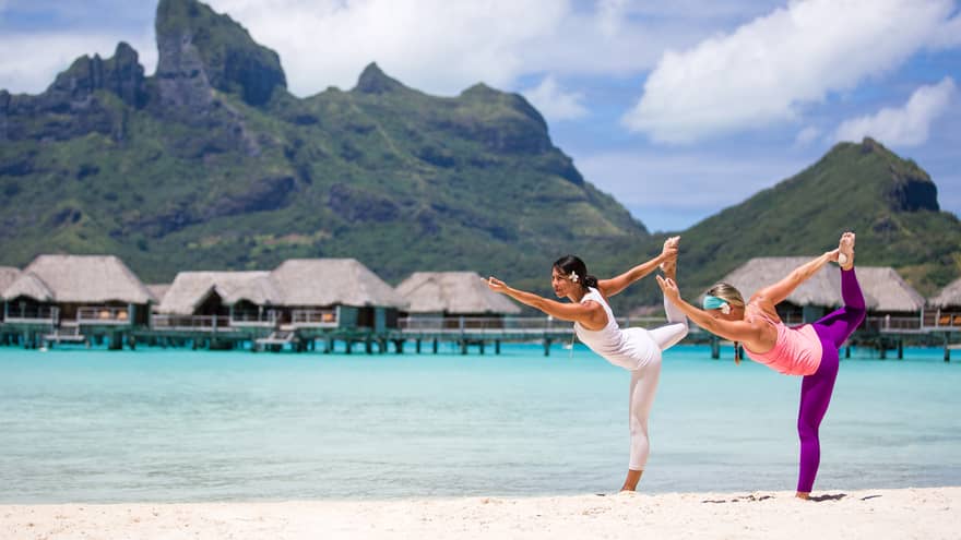 Two people practicing yoga on a tropical beach with overwater bungalows and mountains in the background