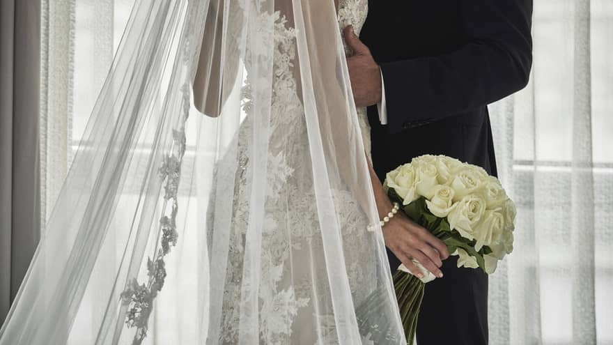 A wedding couple poses in front of a large window, dressed in wedding attire, with a long lace veil, holding a bouquet of white roses