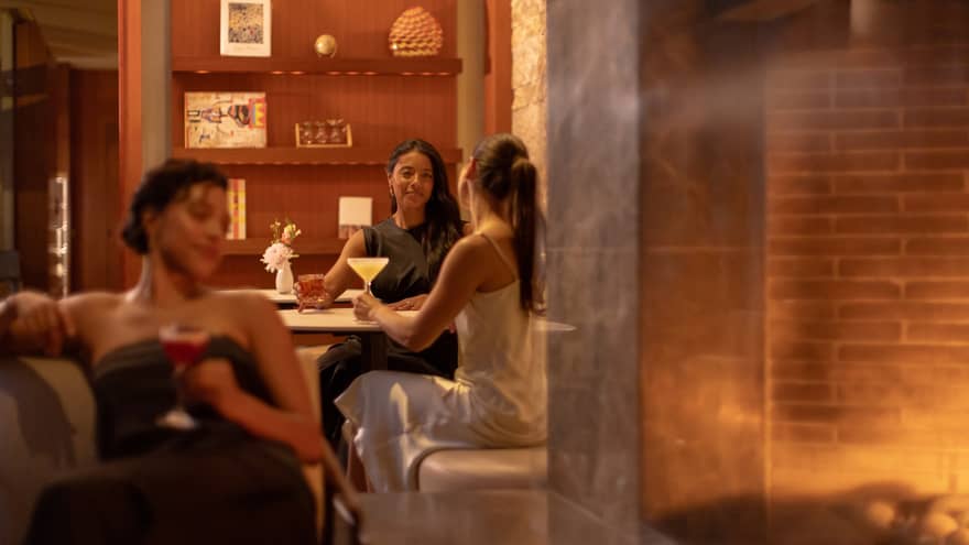 One guest sits on a couch with a drink while two other guests hold their drinks at a dining table