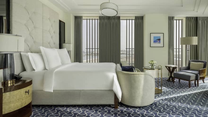 Bright hotel bedroom with a plush white bed, tufted headboard, a sitting area with chairs and a table, and large windows offering views of the city and water