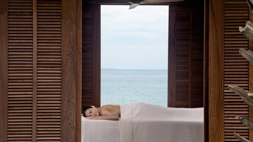 Guest enjoying a relaxing outdoor massage with a serene ocean view, framed by wooden shutters at a luxury beachfront resort
