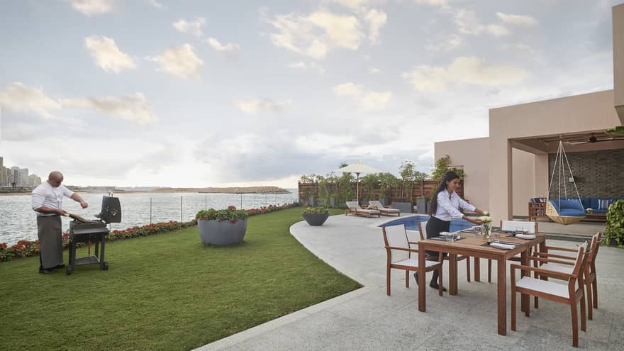 A chef seasons meat on a barbecue near the water’s edge while a server arranges flowers at a table on a patio near a pool.