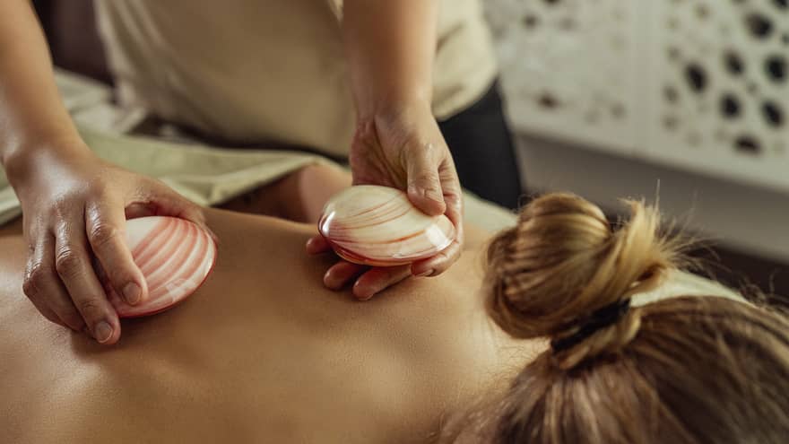Close up of masseuse's hands placing red-and-white shells on the bare back of a person lying face down