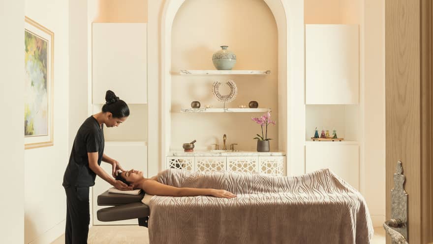 Spa treatment room, staff massages woman's head as she lays under blanket on massage bed 
