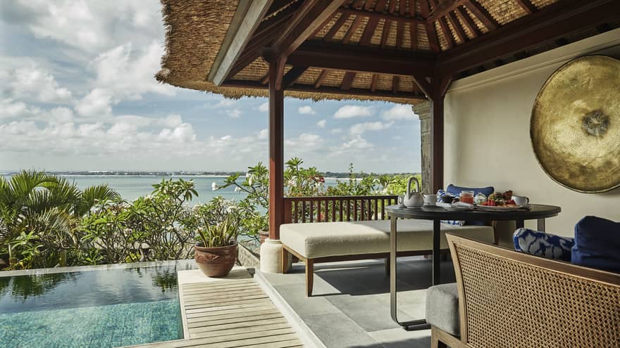 Close-up of Deluxe Villa patio corner with swimming pool, plant, white bench under thatched roof, ocean view