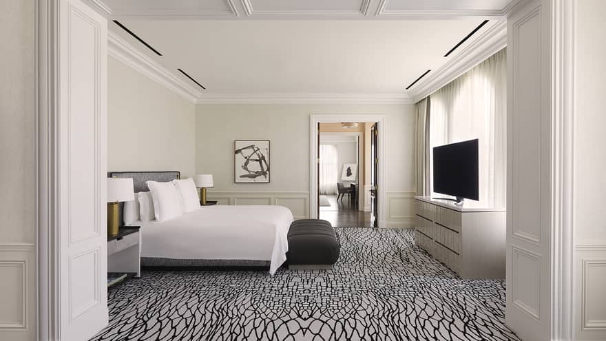 Bedroom with white and black carpeting, white king bed, black bench, TV and dresser