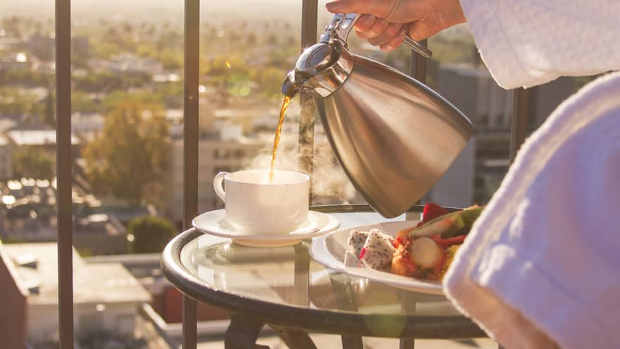 Close up of person in bathrobe pouring coffee from steel carafe, breakfast fruit plate on balcony