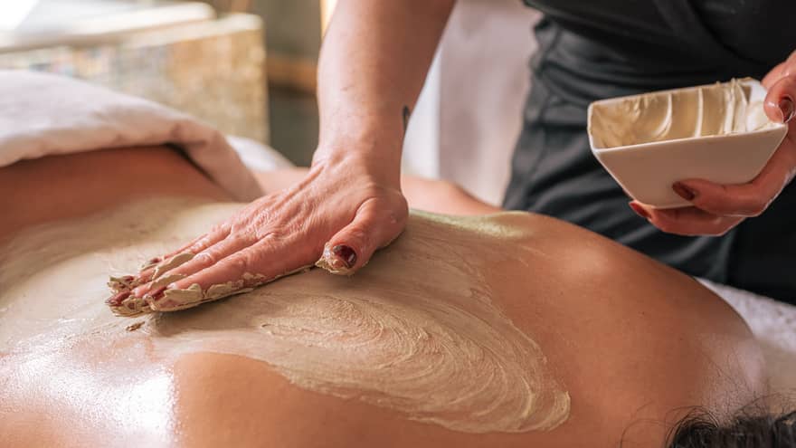 A hand rubs a sand-coloured body treatment on a person's bare back