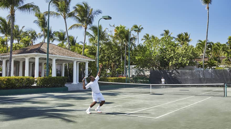A man in all white trows a tennis ball high into the air, mid-serve 
