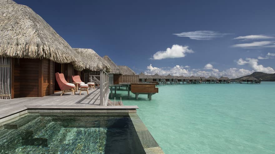 Overwater bungalows with thatched roofs, private pools and lounge chairs overlooking a clear turquoise lagoon under a bright blue sky.