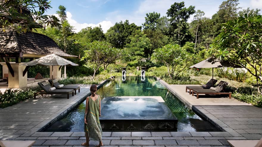 Back of woman in long beach dress walking towards residence swimming pool surrounded by tropical forest