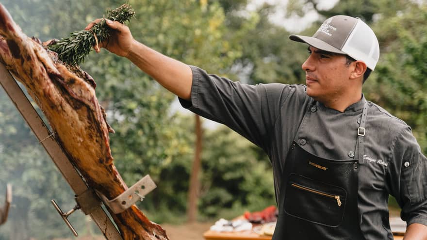 Chef wearing dark grey shirt, black apron and grey-and-white ball cap holds a bundle of herbs in one hand as he swipes it over a large piece of meat hanging on an outdoor grill