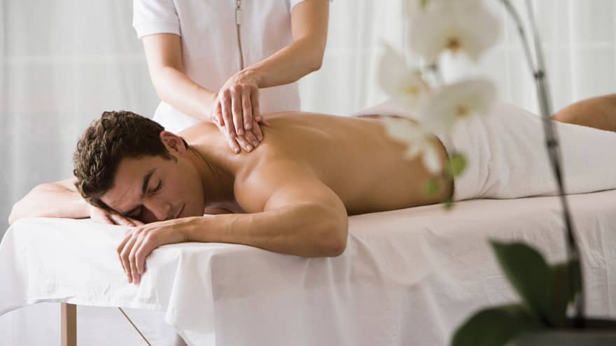 Man lays on white table with eyes closed as masseuse massages his bare shoulders
