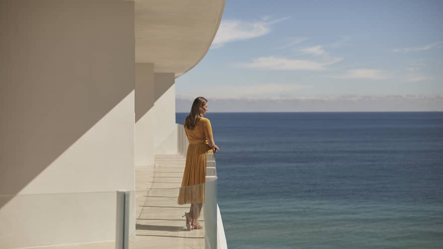 A woman standing on a balcony  of a white stone building with the ocean in the background.