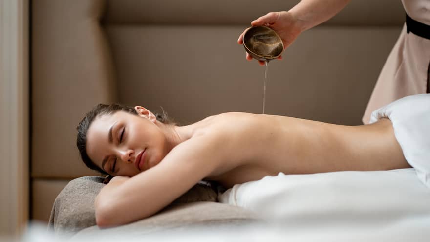 Woman lying with eyes closed on stomach while spa therapist pours oil from dish on her back