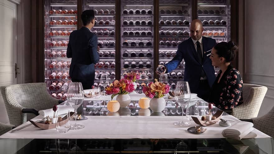 Man scans glass cellar as waitstaff pours wine in glass for woman at table