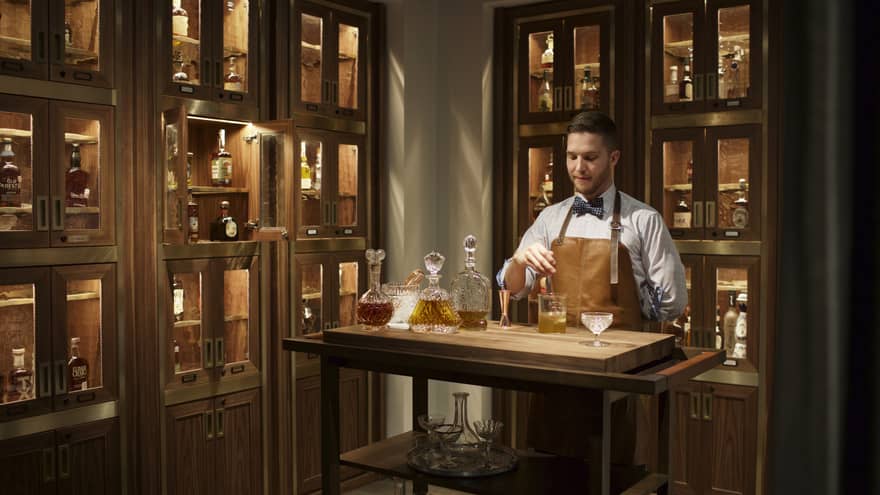 Bayou & Bottle mixologist in apron stirs bourbon cocktail, surrounded by illuminated display cases with bottles 