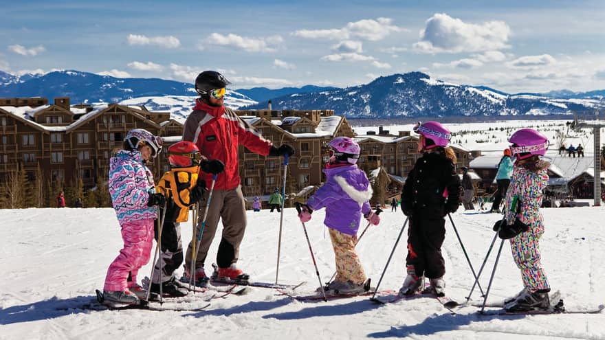 Children wearing skis, winter gear on sunny hill with instructor, hotel in background