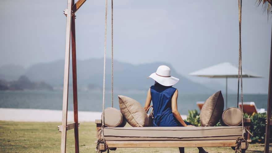 Woman in blue dress and white sun hat lounges on outdoor swing