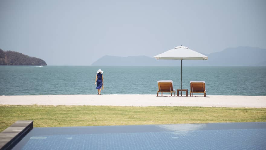 Infinity pool, lawn and beach. A woman walks on the beach next to two lounge chairs and umbrella.