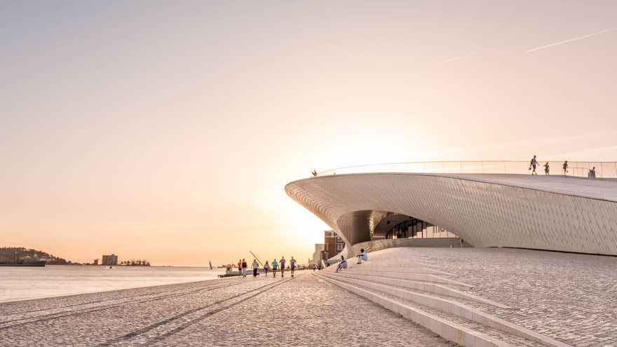 Tourists walk by a disc-shaped modern building next to a harbour. The setting sun reflects off the cobbled walkway. 