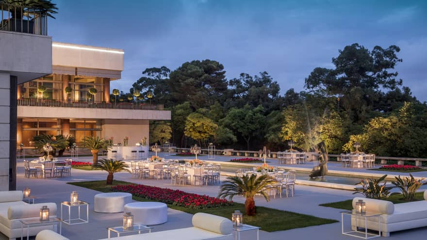 Modern white patio lounge furniture, tables, potted palms surrounding fountain at dusk