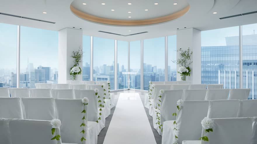 An indoor wedding venue with rows of white chairs and flowers, with a view of the city skyline from window.