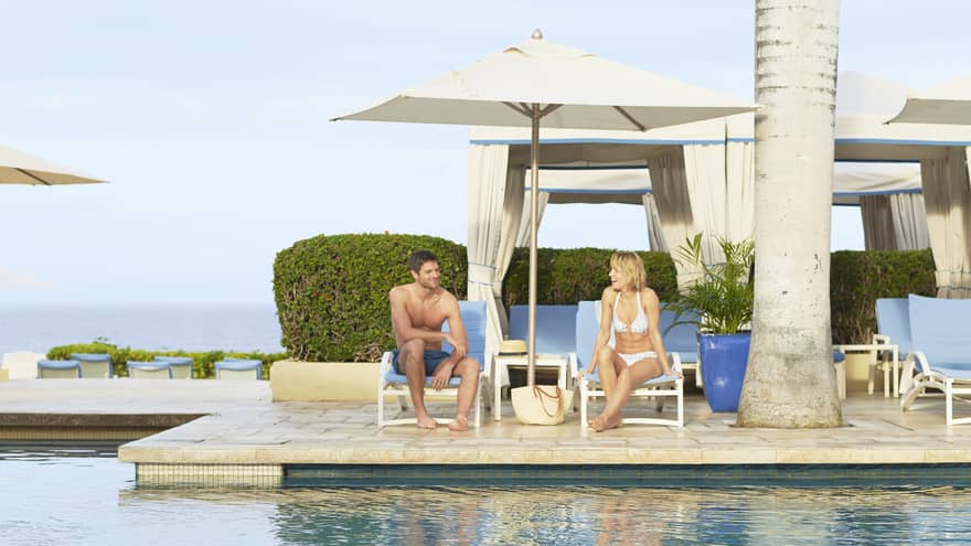Two people relaxing on lounge chairs by the pool, under umbrellas with cabanas and ocean view in the background 
