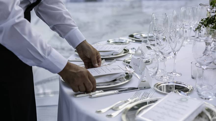 A server in a white shirt sets a formal dining table, placing a menu on a silver plate. The table is elegantly arranged with glassware, silverware and name cards.