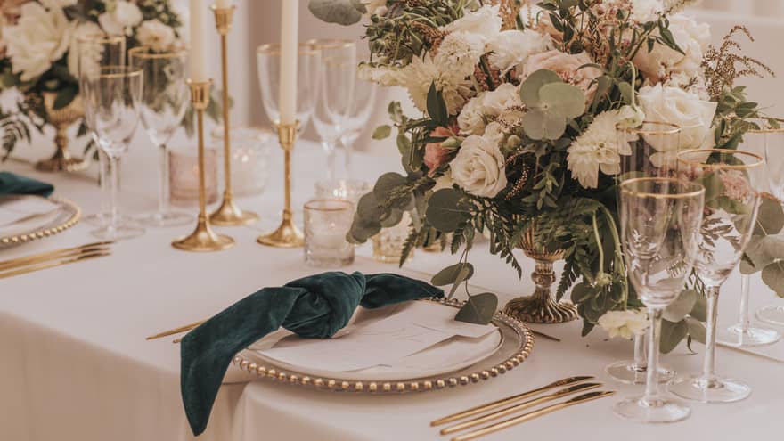 A beautifully arranged table adorned with gold cutlery, white roses and dark green knotted napkins, setting the vibe for a perfect wedding dinner