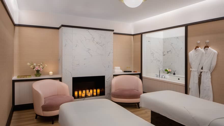 Couple's spa suite with a white-tiled fireplace filled with lit candles, an inset bathtub, two pink arm chairs, two side-by-side treatment tables and a pair of white robes