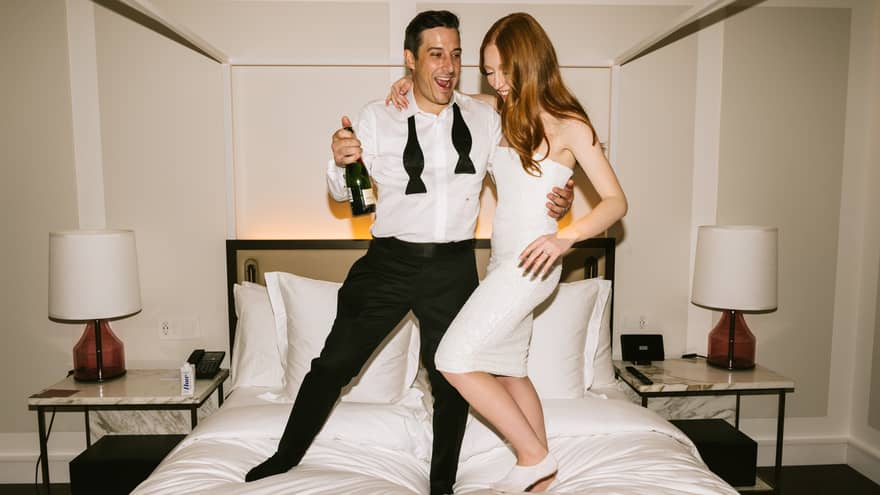 A stylish couple, dressed in an elegant white dress, shirt and black pants, joyfully stand on a bed, celebrating with a bottle of champagne