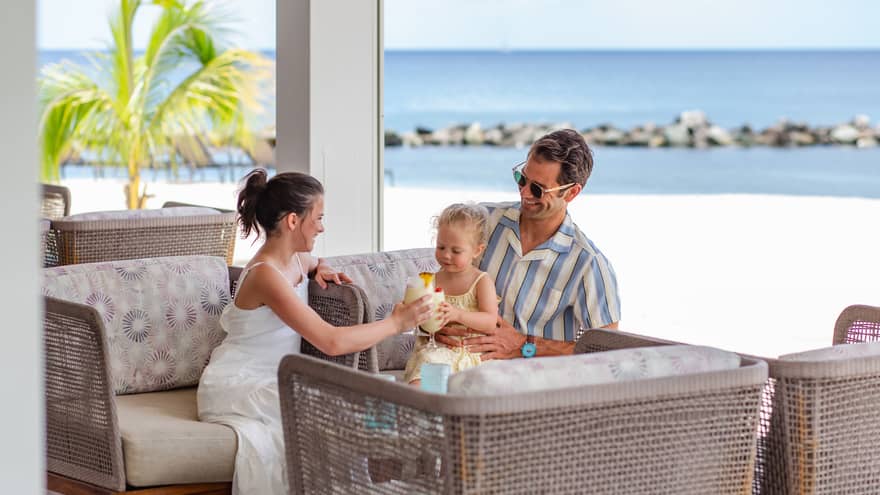 An adult wearing a blue-and-white striped button down short-sleeve shirt holds a young child on his lap, while a young teenager in a white dress sits next to them playing with the baby with the beach in the background
