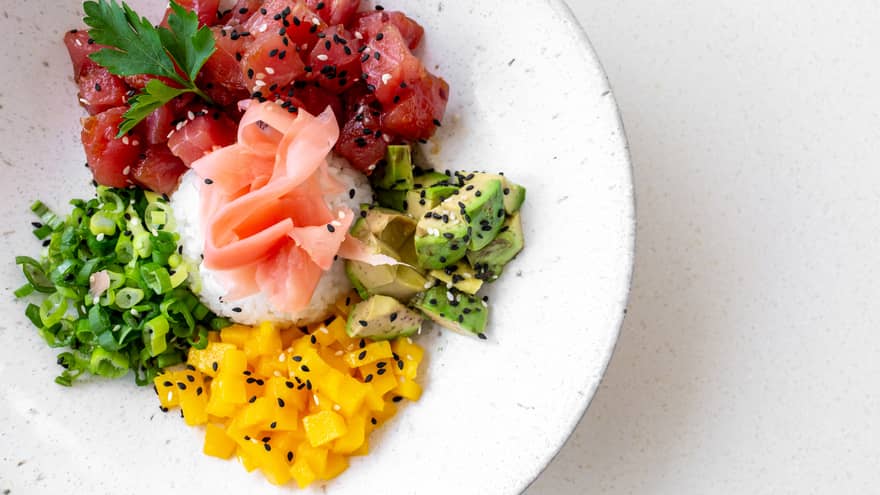 Poke bowl with chunks of tuna, mango, scallions and avocado plated with a rice, cilantro, sesame seeds and orange ponzu