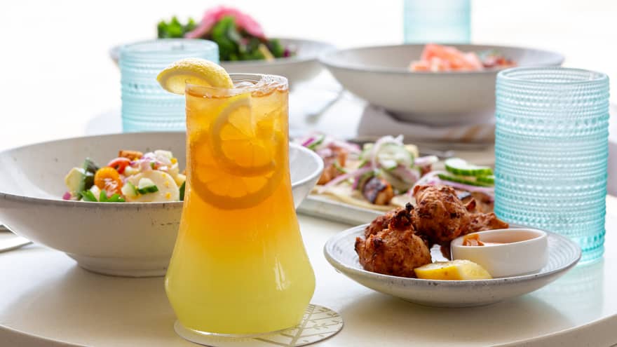 Large glass pitcher of orange juice, a small plate of chicken wings, a bowl of salad, a rectangular plate of tacos and two more bowls of food fill a white table